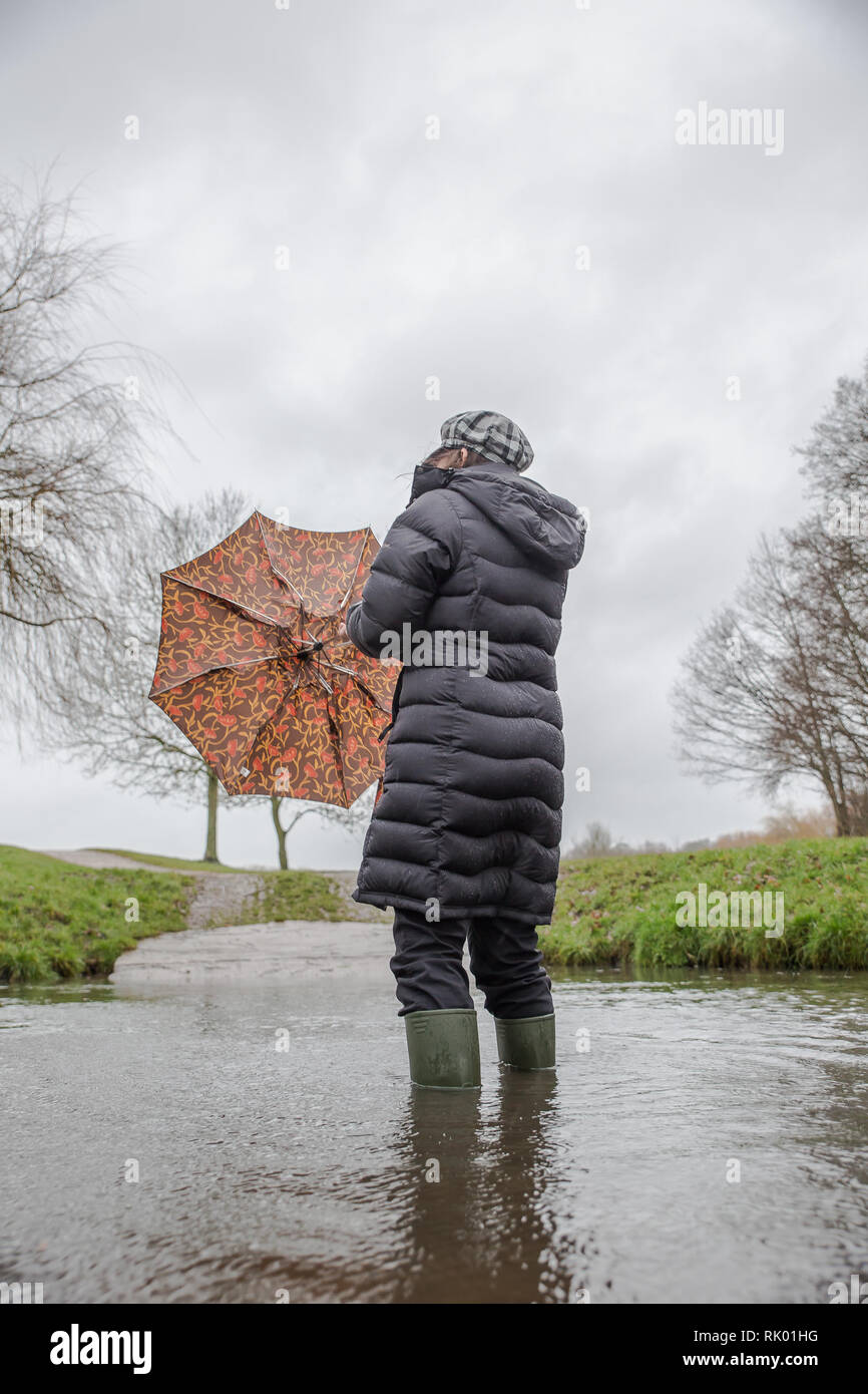 Kidderminster, UK. 8th February, 2019. UK weather: persistent heavy rain leads to water levels rising in Worcestershire with blustery showers continuing throughout the day. A young lady, rear view, wearing a long, winter coat and wellies stands isolated outdoors in water, struggling with her umbrella in the wet and windy conditions. Credit: Lee Hudson/Alamy Live News Stock Photo