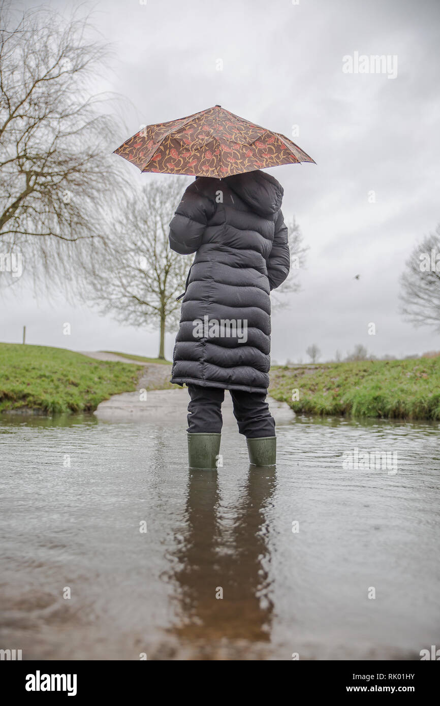 Kidderminster, UK. 8th February, 2019. UK weather: persistent heavy rain leads to water levels rising in Worcestershire with blustery showers continuing throughout the day. A young lady, rear view, wearing a long, winter coat and wellies stands isolated outdoors in water, struggling with her umbrella in the wet and windy conditions. Credit: Lee Hudson/17�Թ����� Live News Stock Photo