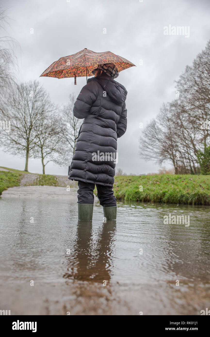 Kidderminster, UK. 8th February, 2019. UK weather: persistent heavy rain leads to water levels rising in Worcestershire with blustery showers continuing throughout the day. A young lady, rear view, wearing a long, winter coat and wellies stands isolated outdoors in water, struggling with her umbrella in the wet and windy conditions. Credit: Lee Hudson/Alamy Live News Stock Photo