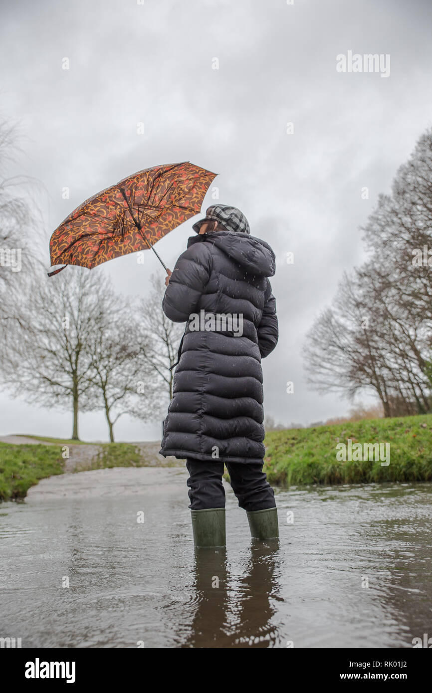 Kidderminster, UK. 8th February, 2019. UK weather: persistent heavy rain leads to water levels rising in Worcestershire with blustery showers continuing throughout the day. A young lady, rear view, wearing a long, winter coat and wellies stands isolated outdoors in water, struggling with her umbrella in the wet and windy conditions. Credit: Lee Hudson/Alamy Live News Stock Photo