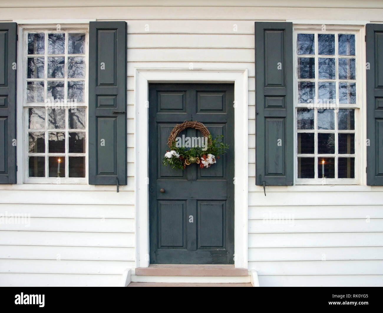 white frame house, dark green shutters & door, grapevine Christmas wreath, cotton bolls; evergreens, lighted candles; Colonial Williamsburg; VA; Virgi Stock Photo