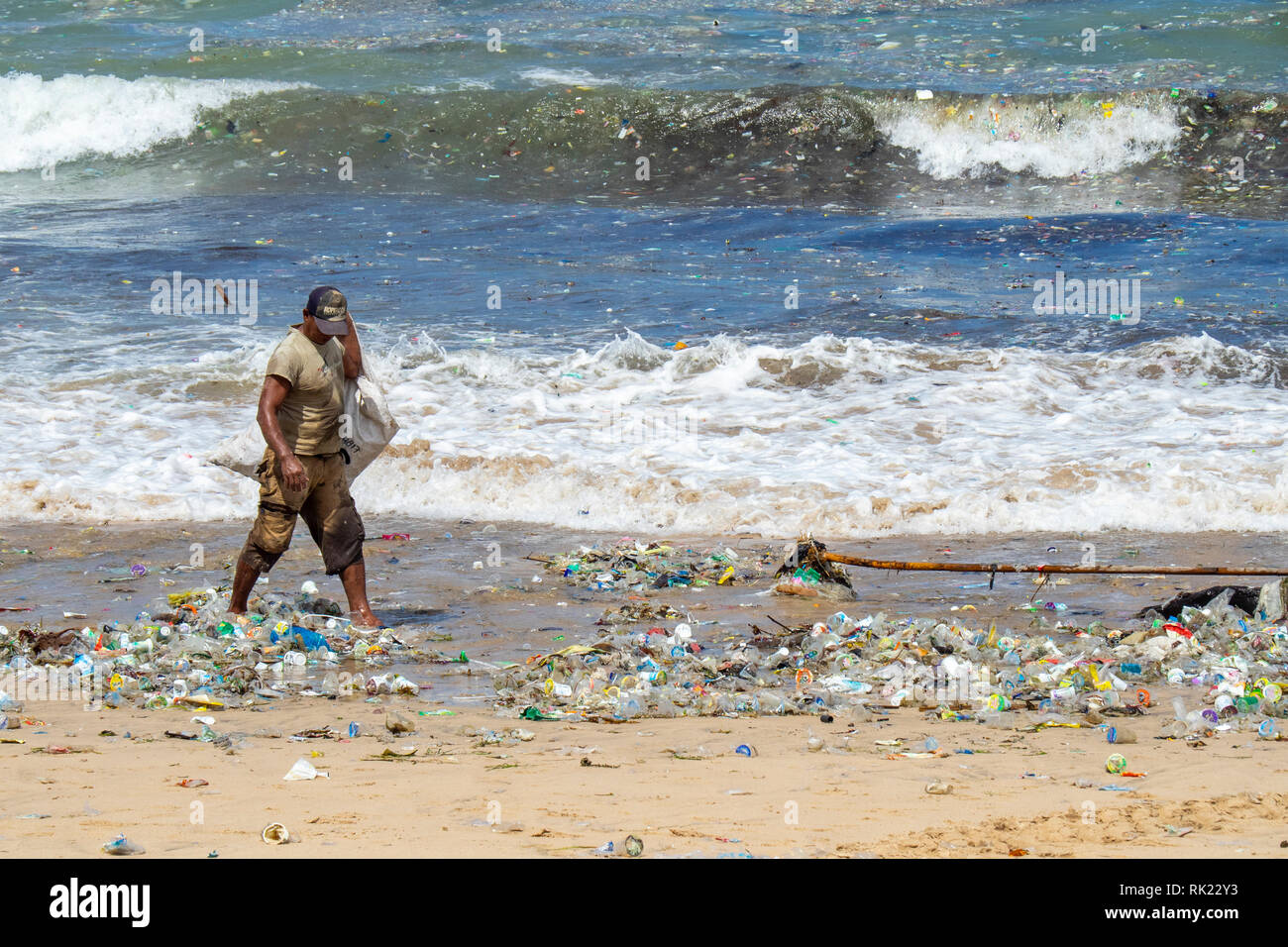 Pollution, lone man picking up plastic bottles, cups, straws and other litter washed up on the beach at Jimbaran Bay, Bali Indonesia.. Stock Photo