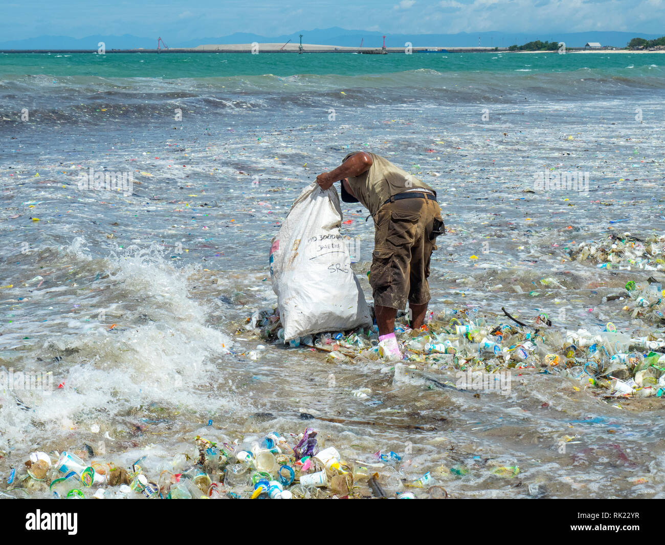 Pollution, lone man picking up plastic bottles, cups, straws and other litter washed up on the beach at Jimbaran Bay, Bali Indonesia.. Stock Photo