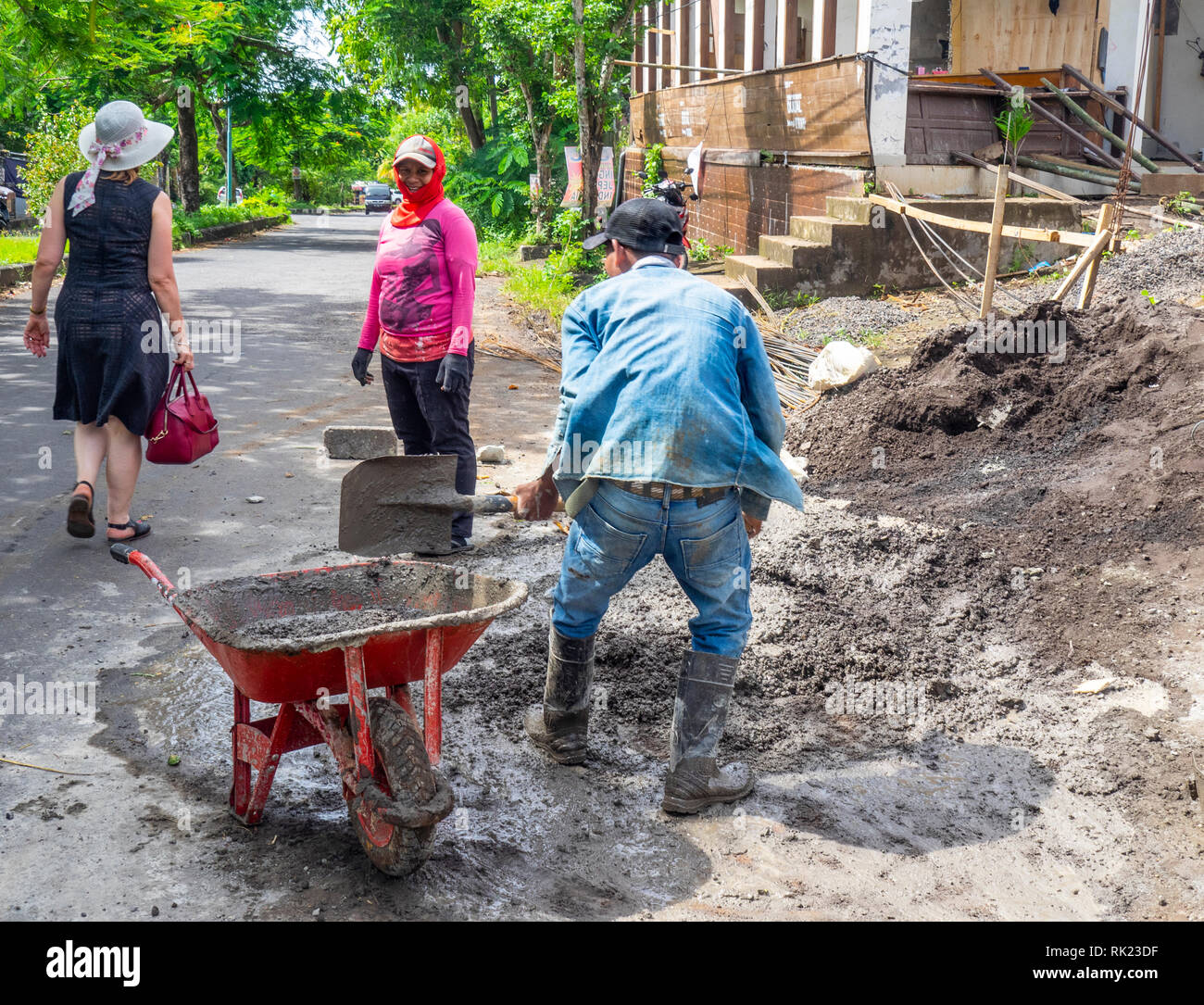 Caucasian tourist walking past a man and a woman workers shovelling cement into a wheelbarrow Jimbaran, Bali Indonesia. Stock Photo