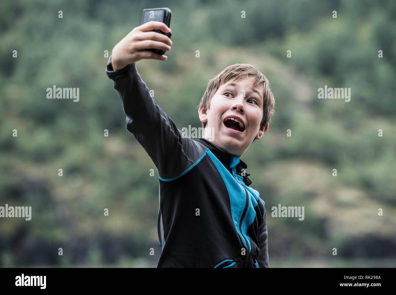 Candid portrait of young boy taking selfie using mobile phone, forest in background, close up Stock Photo