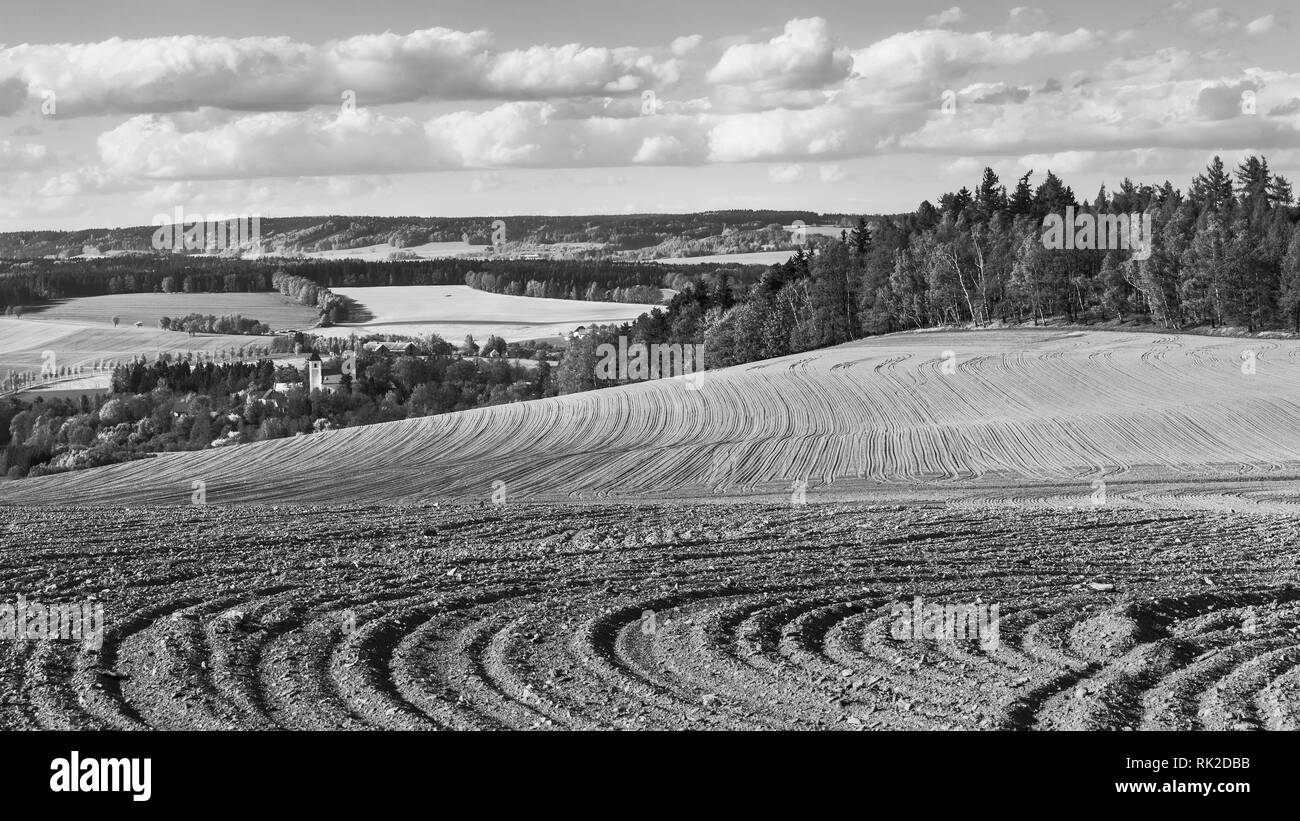 Farmland and forests in black and white landscape. Sown soil close-up. Furrows in sloping field. Spring sky with clouds. Church tower below the hill. Stock Photo