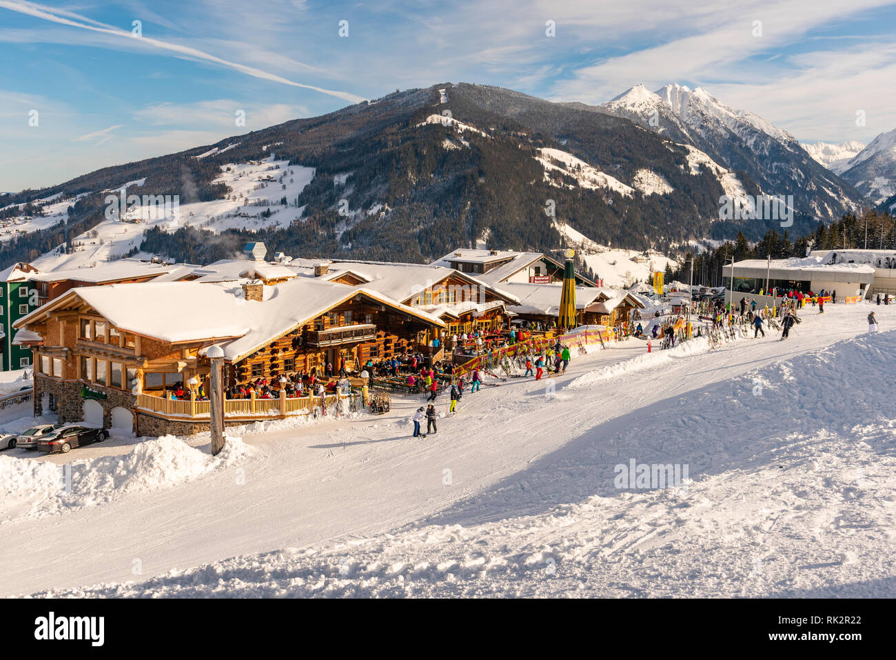 Alpine restaurant full of skiers, snowboarders and tobogganers on sunny day. In the background Hochwurzen gondola ski station, snow-capped mountains. Stock Photo