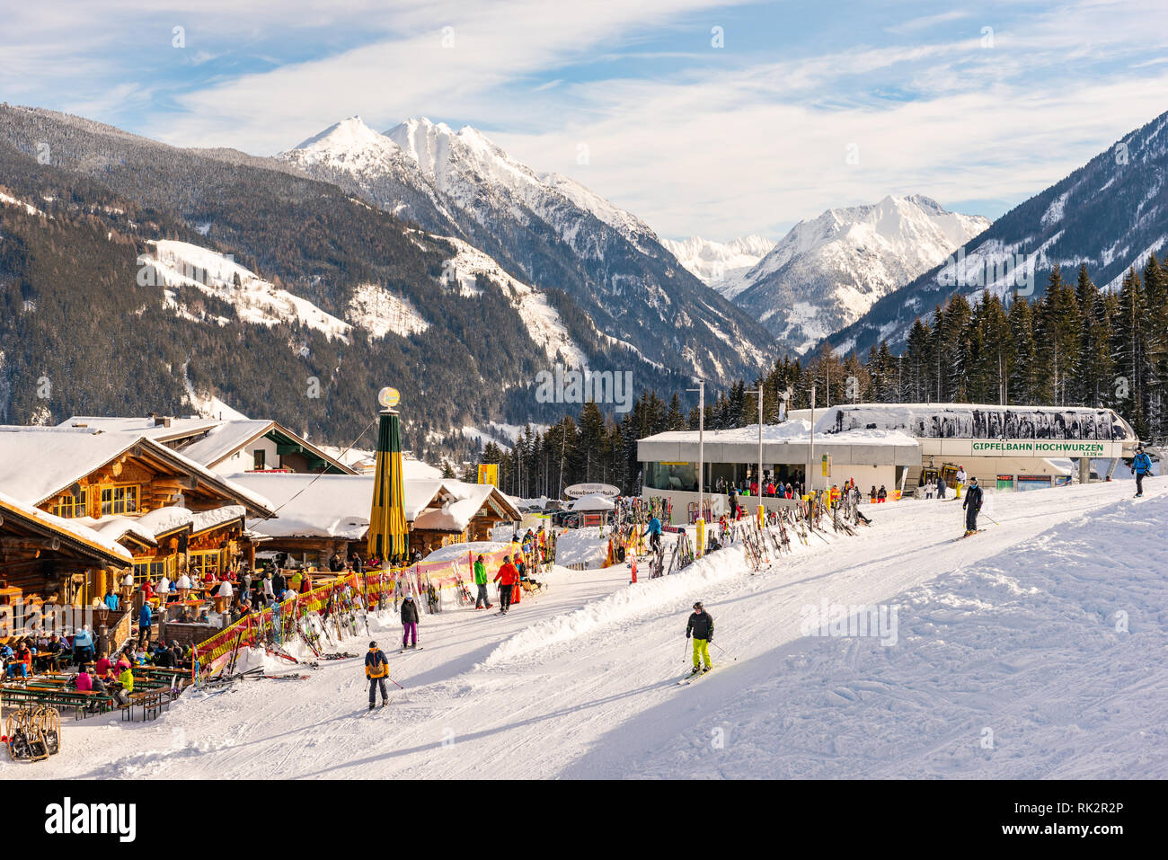 Alpine restaurant full of skiers, snowboarders and tobogganers on sunny day. In the background Hochwurzen gondola ski station, snow-capped mountains. Stock Photo