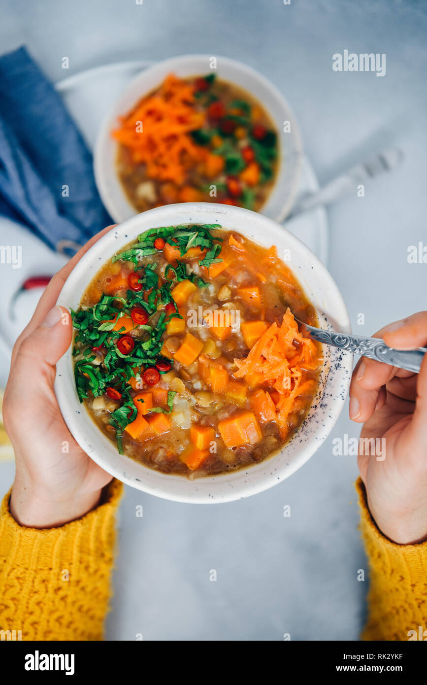 A woman with yellow cardigan holding a bowl of vegan carrot soup with rice, lentils and celeriac, garnished with mint and chili peppers. Stock Photo