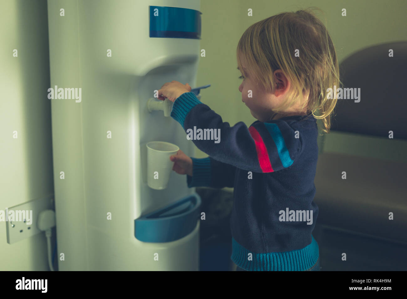 A little toddler is getting a glass of water from the water cooler