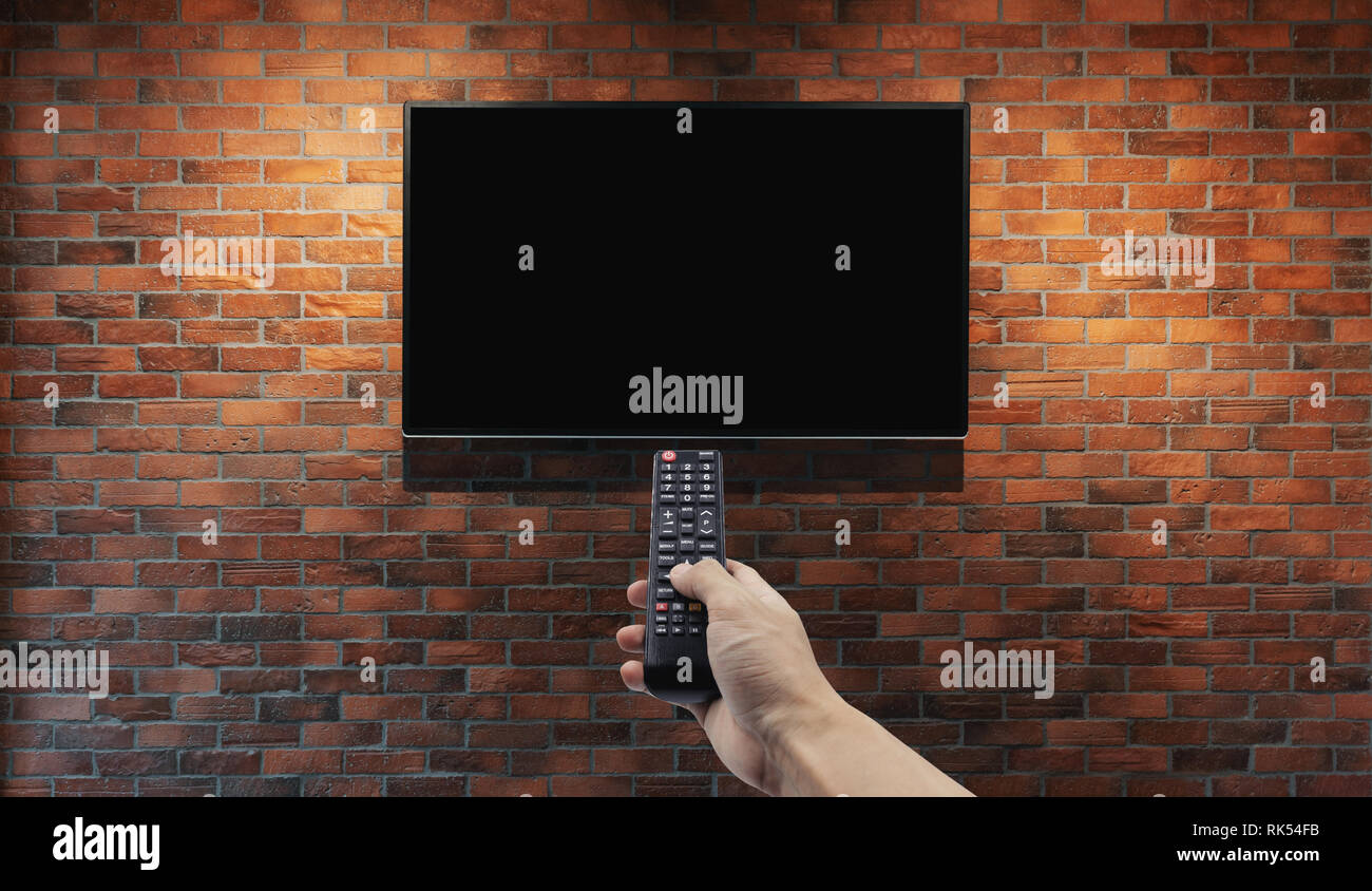 Television on brick wall with hand using remote control Stock Photo
