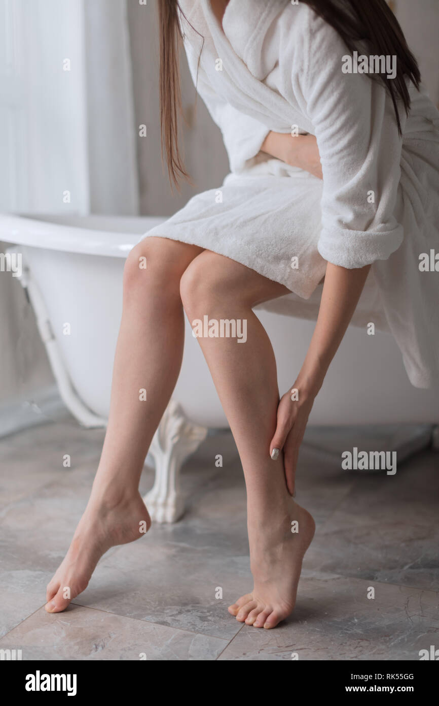 Gorgeous long slim beautiful legs. Close up of young woman after bath, leaning on the edge of bathtub and touching her depilated legs. Hair removal or Stock Photo