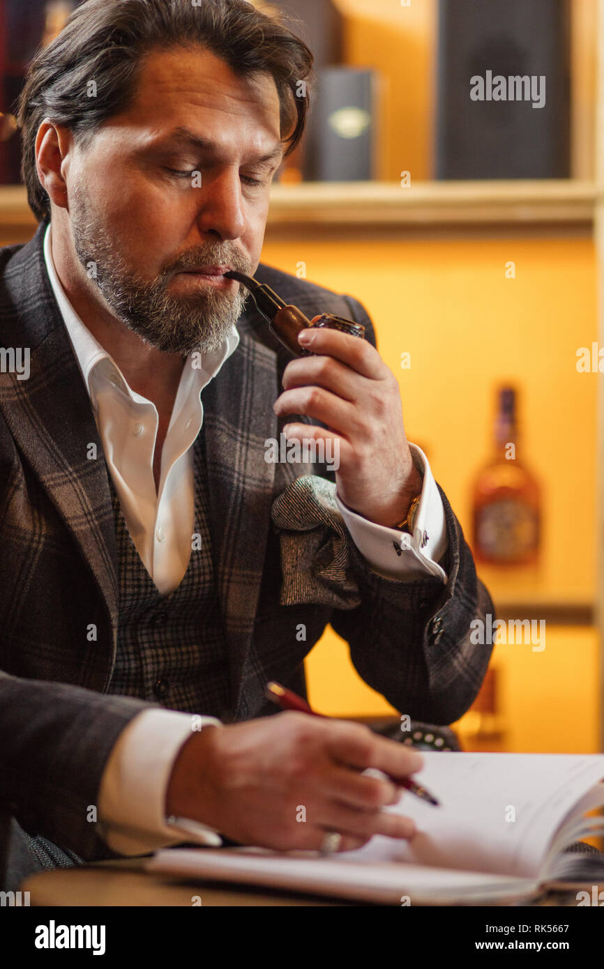 Talanted bearded writer in fashionable clothing is making notes in diary or notebook, sitting at restaurant and enjoying tobacco pipe Stock Photo