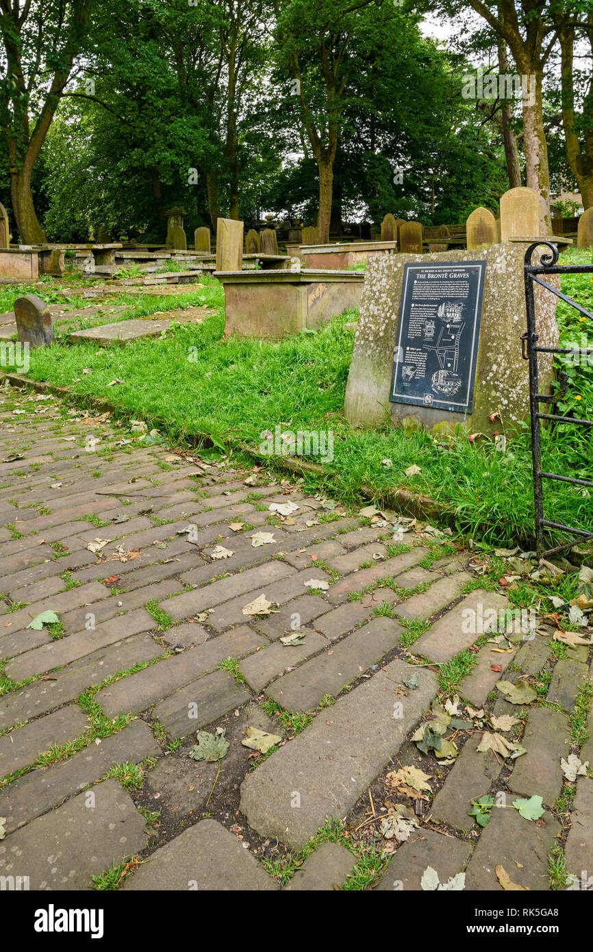 Cobbled path, information plaque (Brontes) headstones & graves in churchyard - St Michael & All Angels Church, Haworth, West Yorkshire, England, UK. Stock Photo