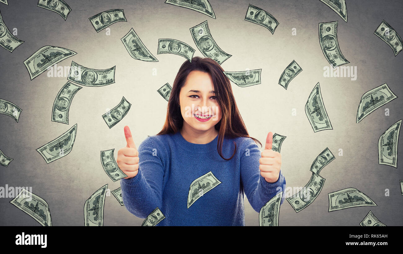 Positive surprised woman showing double thumbs up as money are falling like a rain of dollars from the sky. Lottery winning, celebrating success. Symb Stock Photo
