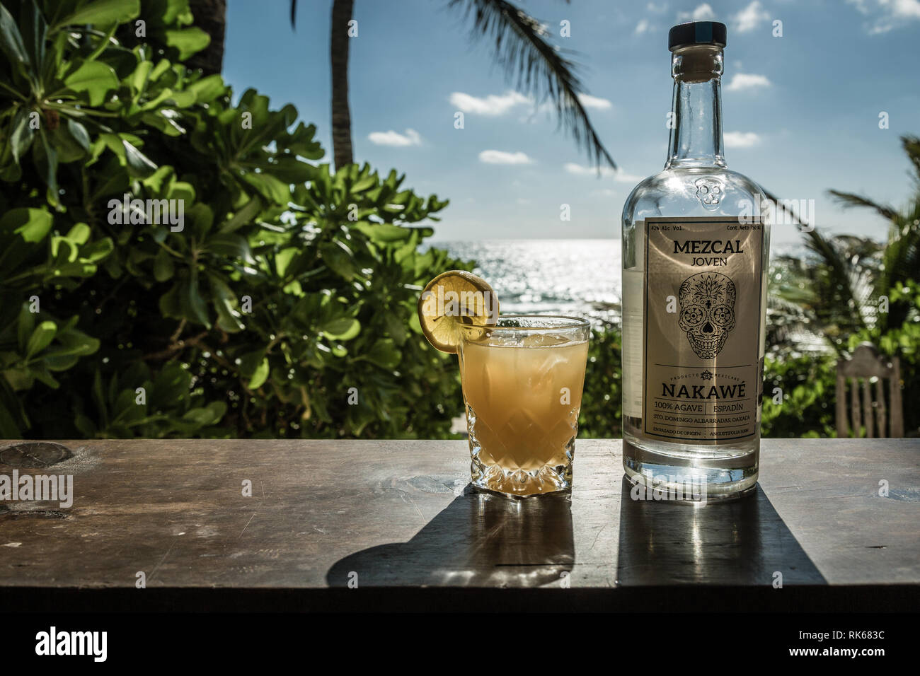 a bottle of mezcal with a glass of orange juice and mezcal shot on a beach bar in tulum with the sea on the background Stock Photo
