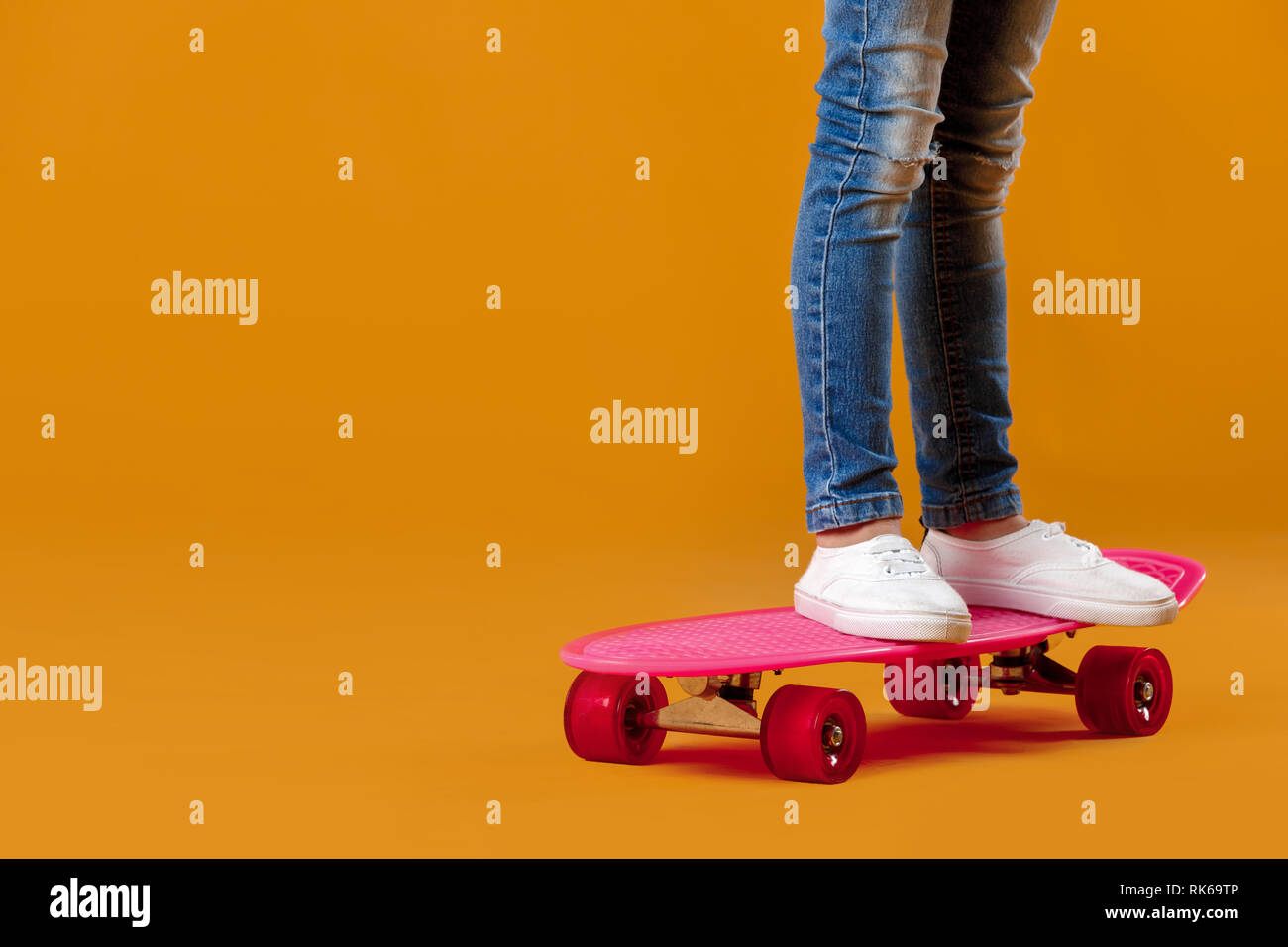 legs of little girl in white sneakers and jeans on pink skateboard on orange background Stock