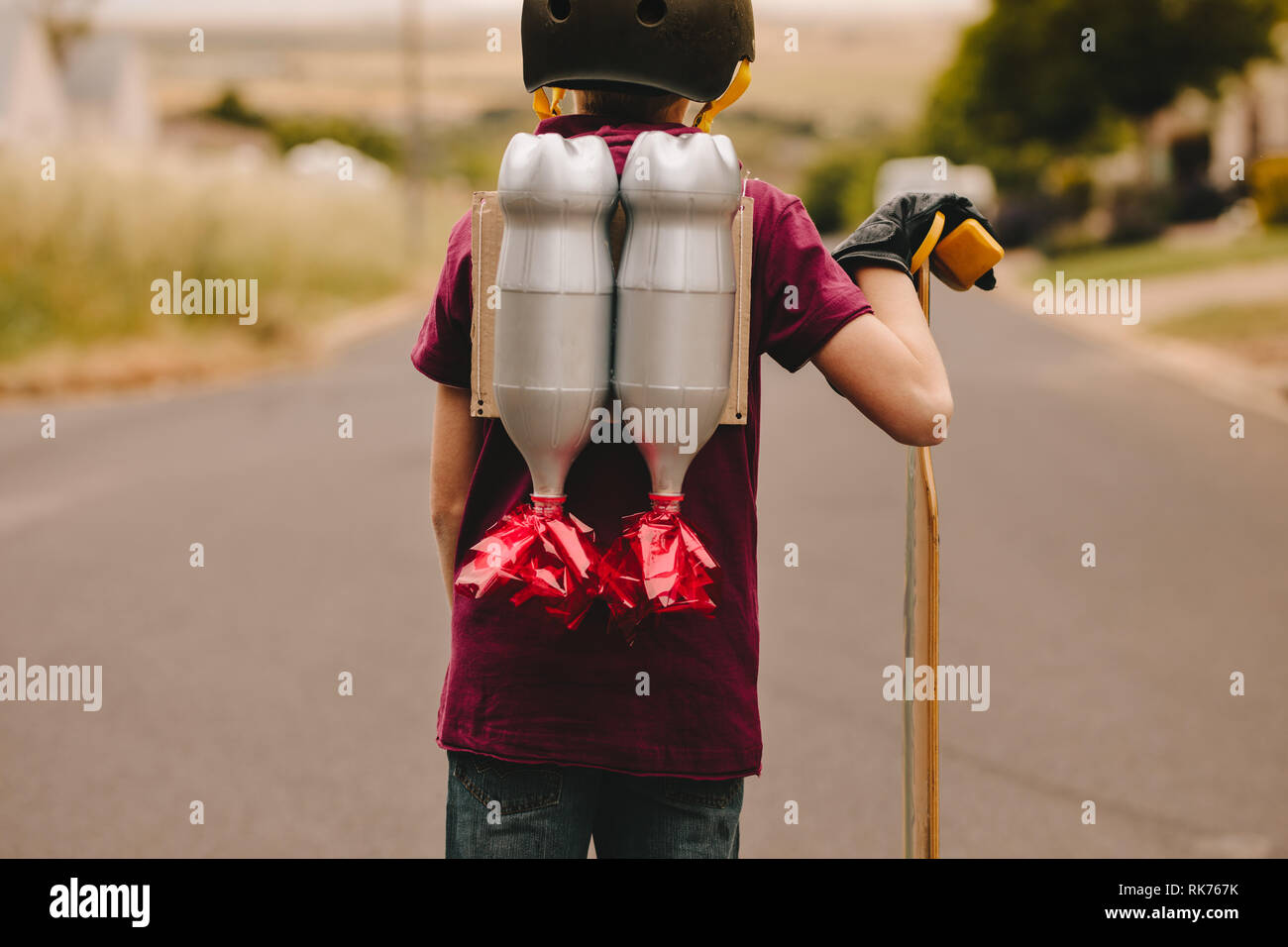 Rear view of young boy with helmet and toy jetpack standing with his skateboard outdoors. Kid with jetpack and skateboard. Stock Photo