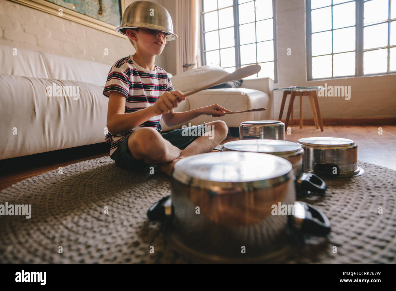Boy playing drums on kitchenware at home. Boy pretending to be a drummer sitting on the living room floor and playing on utensils while wearing a bowl Stock Photo