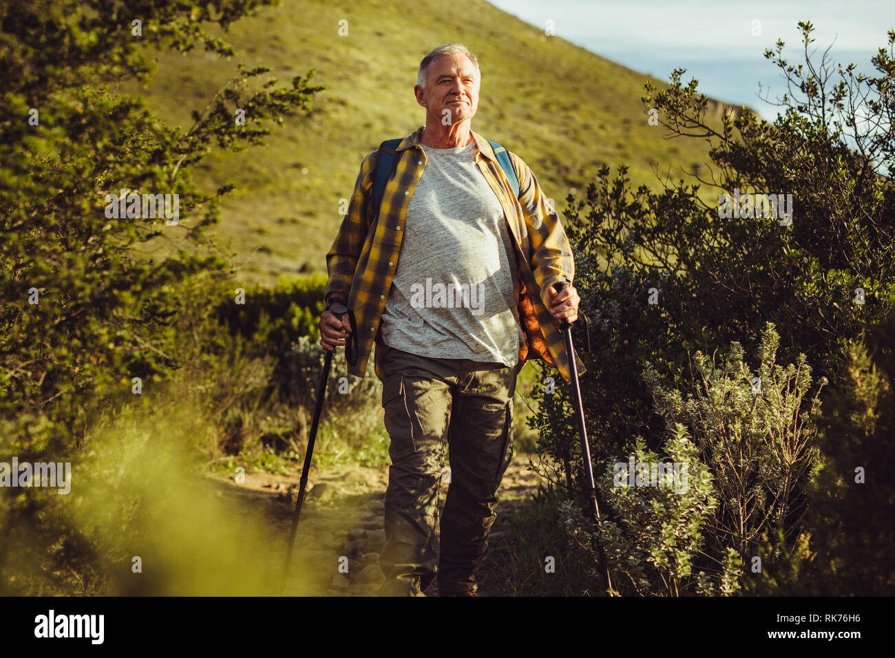 Adventurous man on a trekking trip on a hilly terrain. Senior man walking on a hilly trail path holding trekking poles. Stock Photo