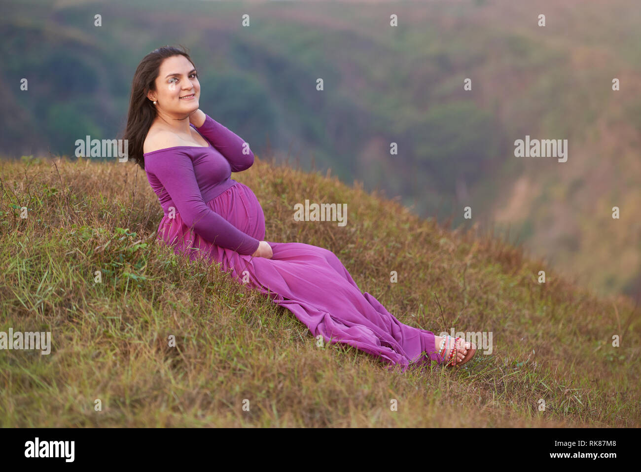 Pregnant woman in purple dress sitting on green field Stock Photo