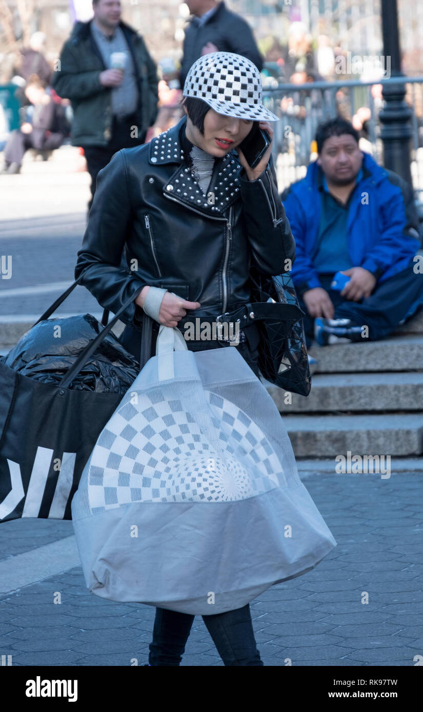 An Asian woman on her cell phone wearing a checkerboard hat and carrying another in a large bag. In Union Square Park in Manhattan, New York City. Stock Photo