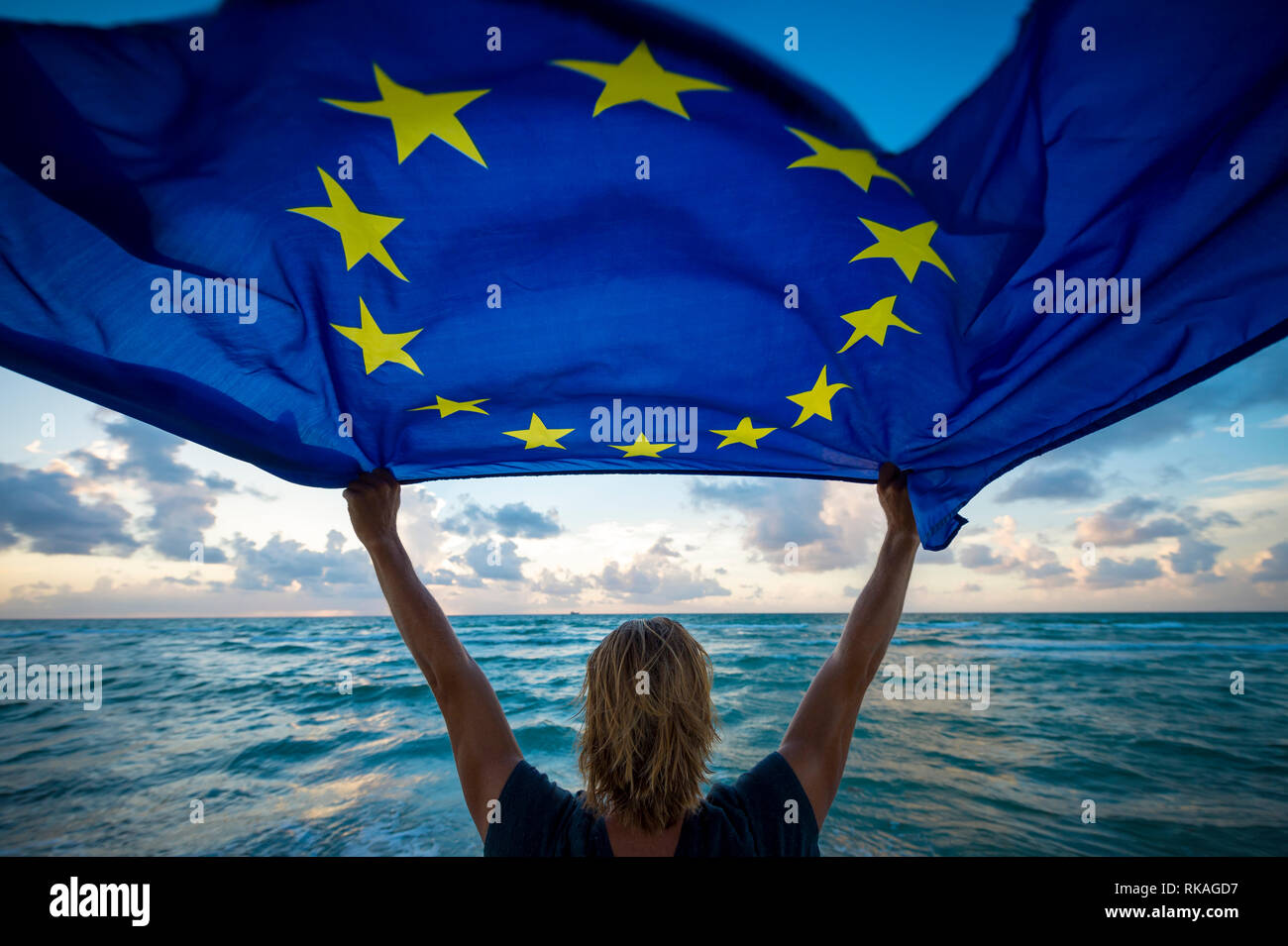 Man holding a fluttering European Union flag flying in bright morning sunlight held up by man with blond hair standing on empty Mediterranean beach Stock Photo