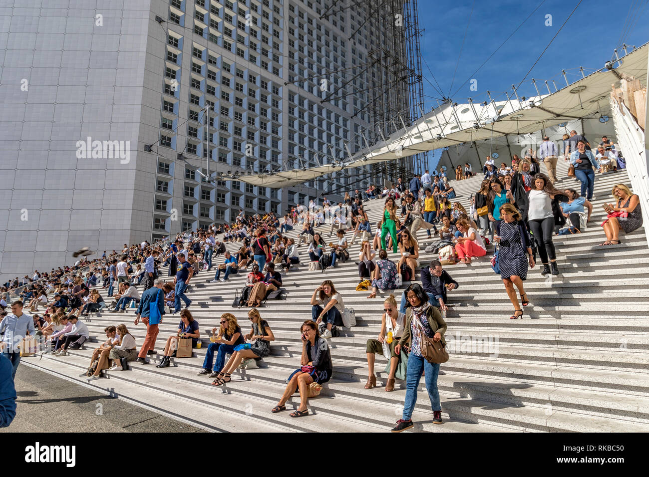 Crowds of people sitting on the steps of La Grande Arche de la Défense enjoying the warm sunshine on a hot summer's day in La Défense ,Paris,France Stock Photo
