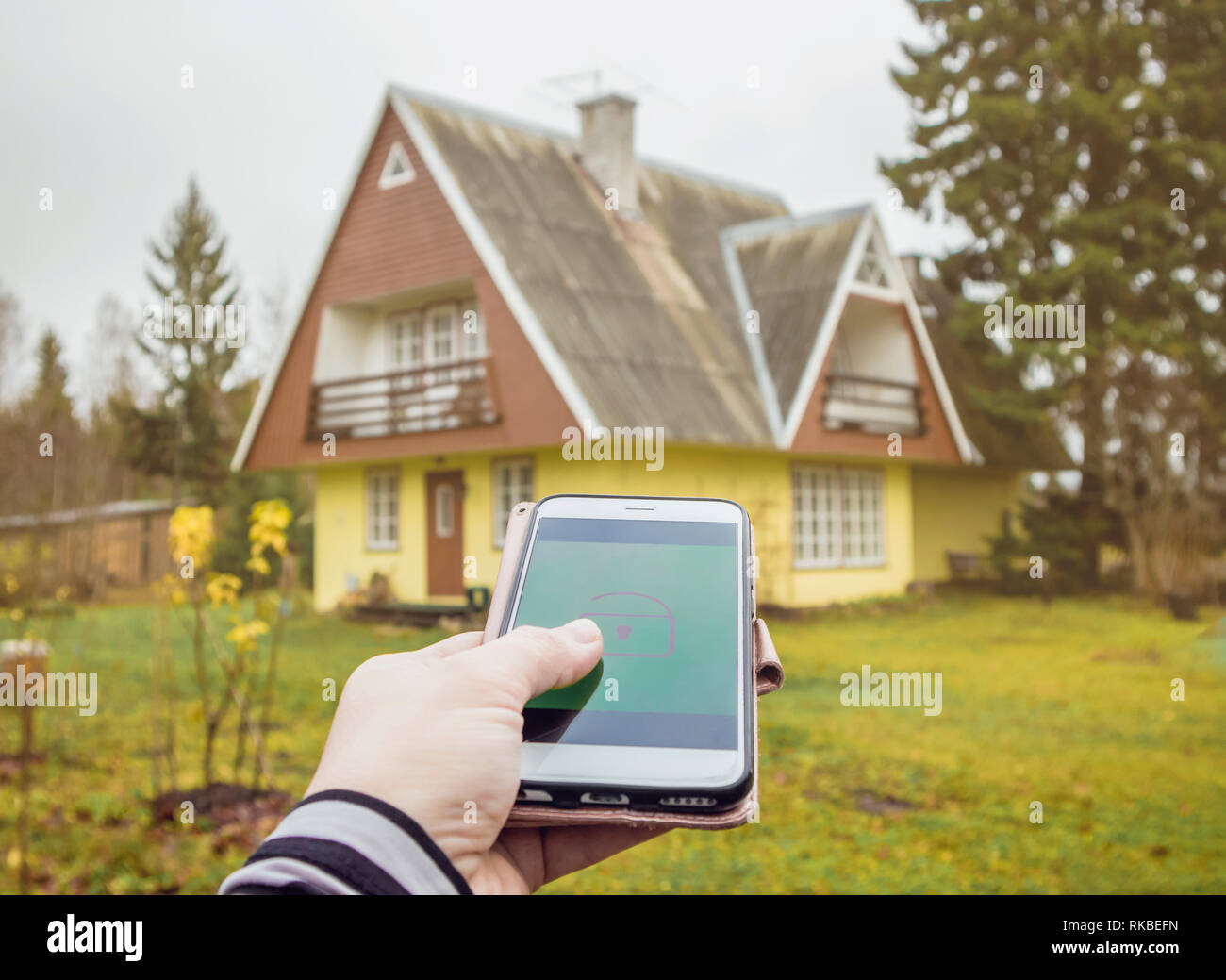 Person hand press lock/unlock button from distance on smartphone app to open/close rural home house door. Modern lifestyle technology concept. Stock Photo