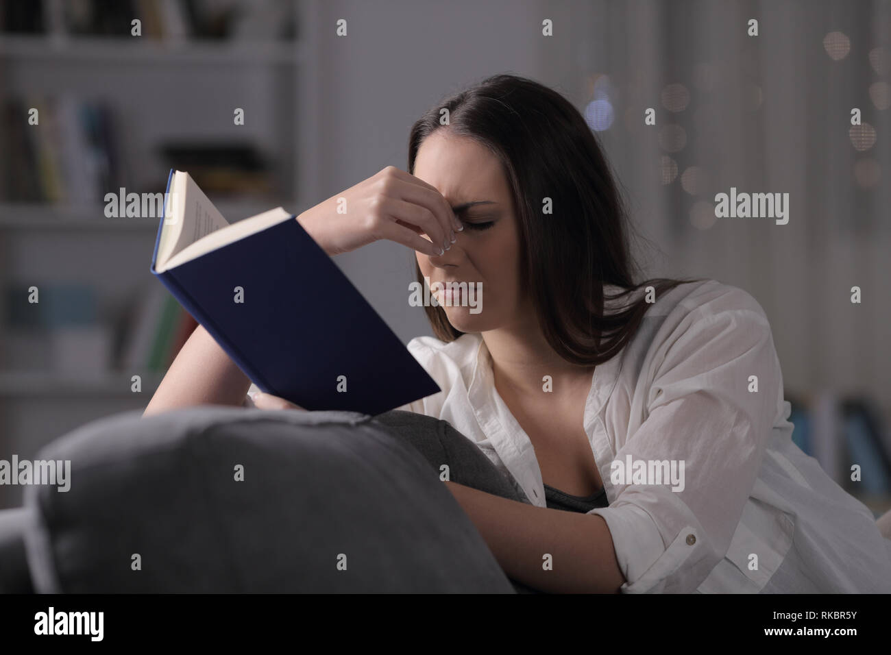 Lady suffering eyestrain reading book sitting on a couch in the night at home Stock Photo