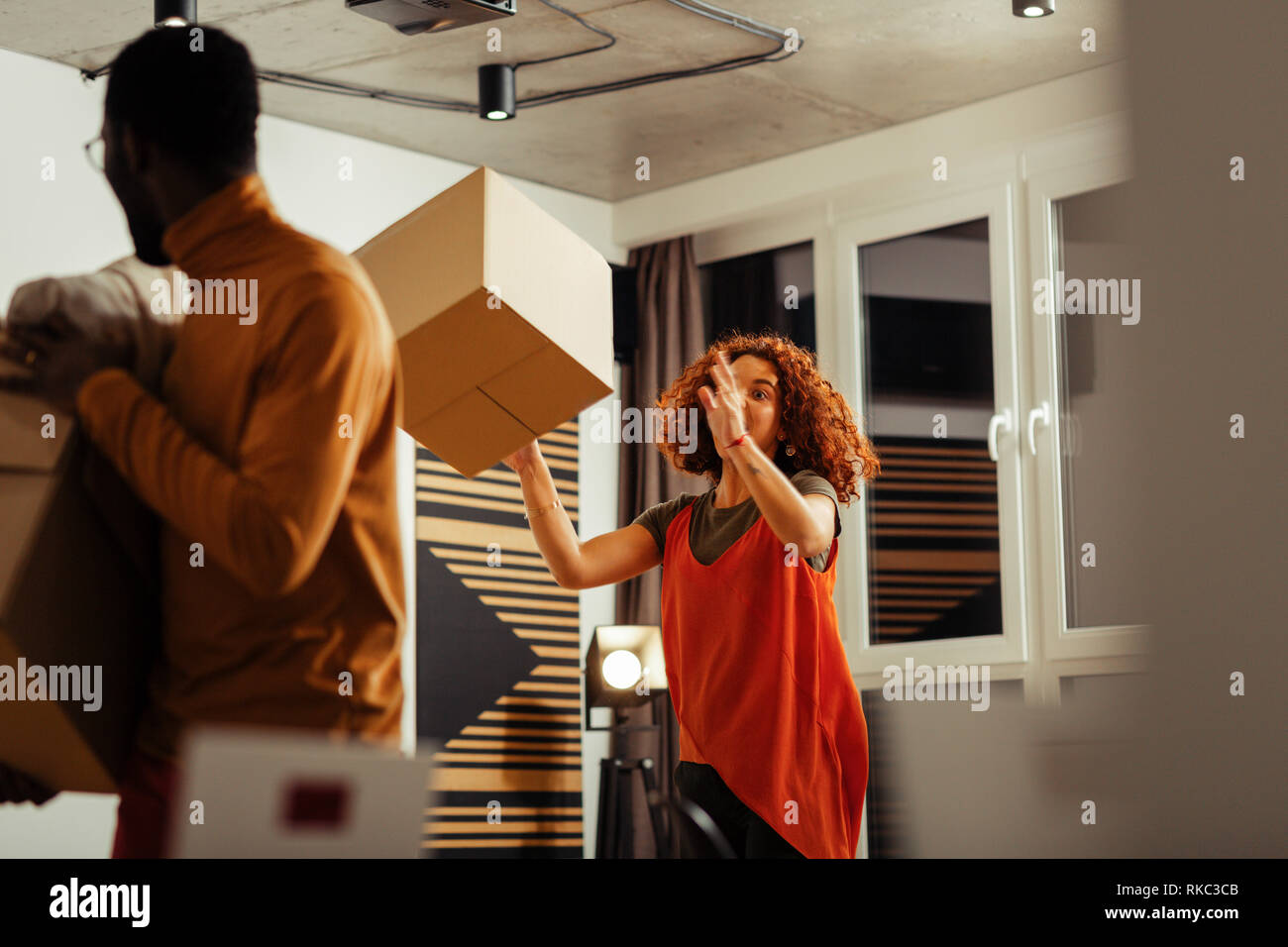Red-haired curly woman lifting box with clothes while moving Stock Photo
