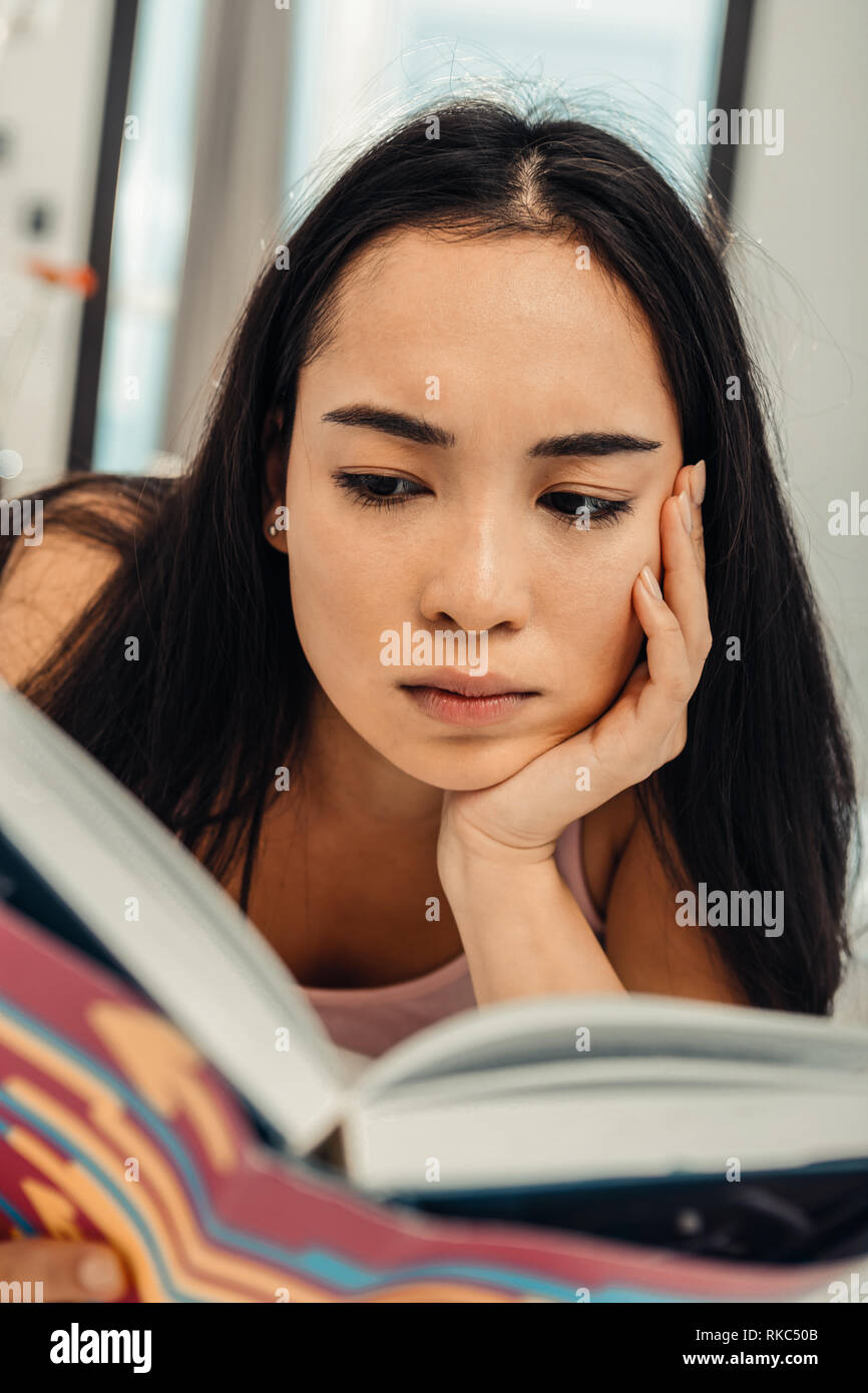 Dark-eyed Asian woman feeling involved in reading book Stock Photo