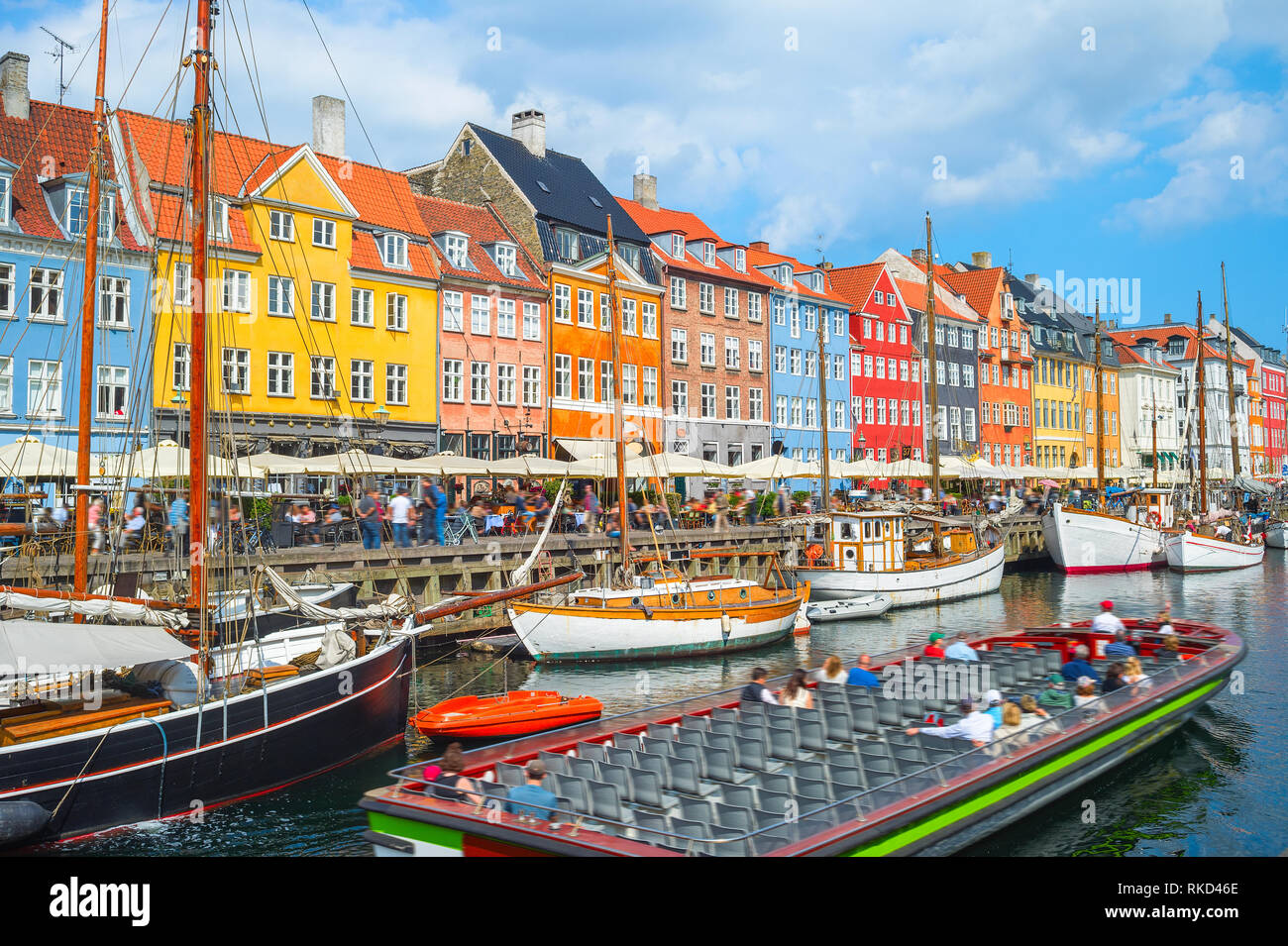Tour boat sailing by canal with old sailboats in Nyhavn harbor, peopel walking at sunny embankment and sitting in cafes and restaurants, Copenhagen, D Stock Photo