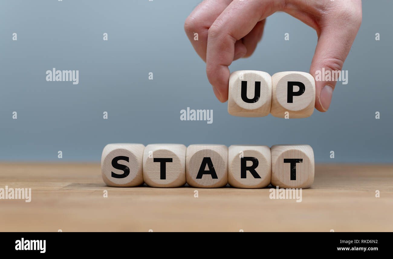 Cubes form the words 'START UP' while to fingers lift the letters 'UP' in the air. Cubes are on a wooden table in-front a grey background. Stock Photo