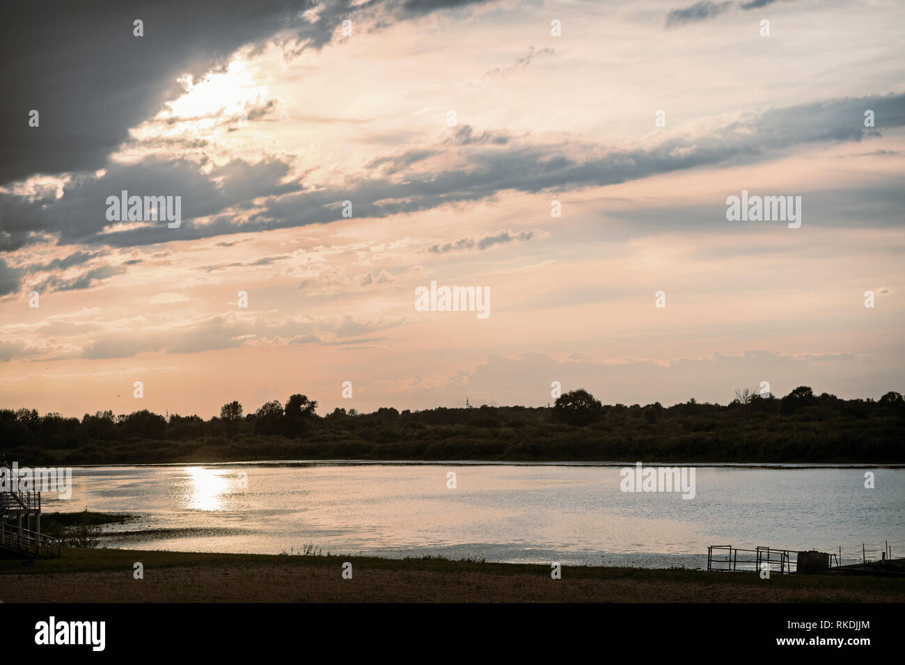 A small river with a wooden Pier is illuminated by the setting sun. The sky is dark and stormy. Stock Photo