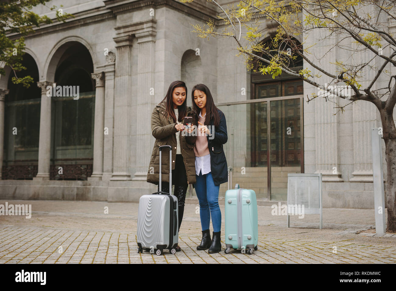 Two asian women tourists looking at mobile phone for navigation. Female tourists standing in street with luggage bags looking for directions in cell p Stock Photo