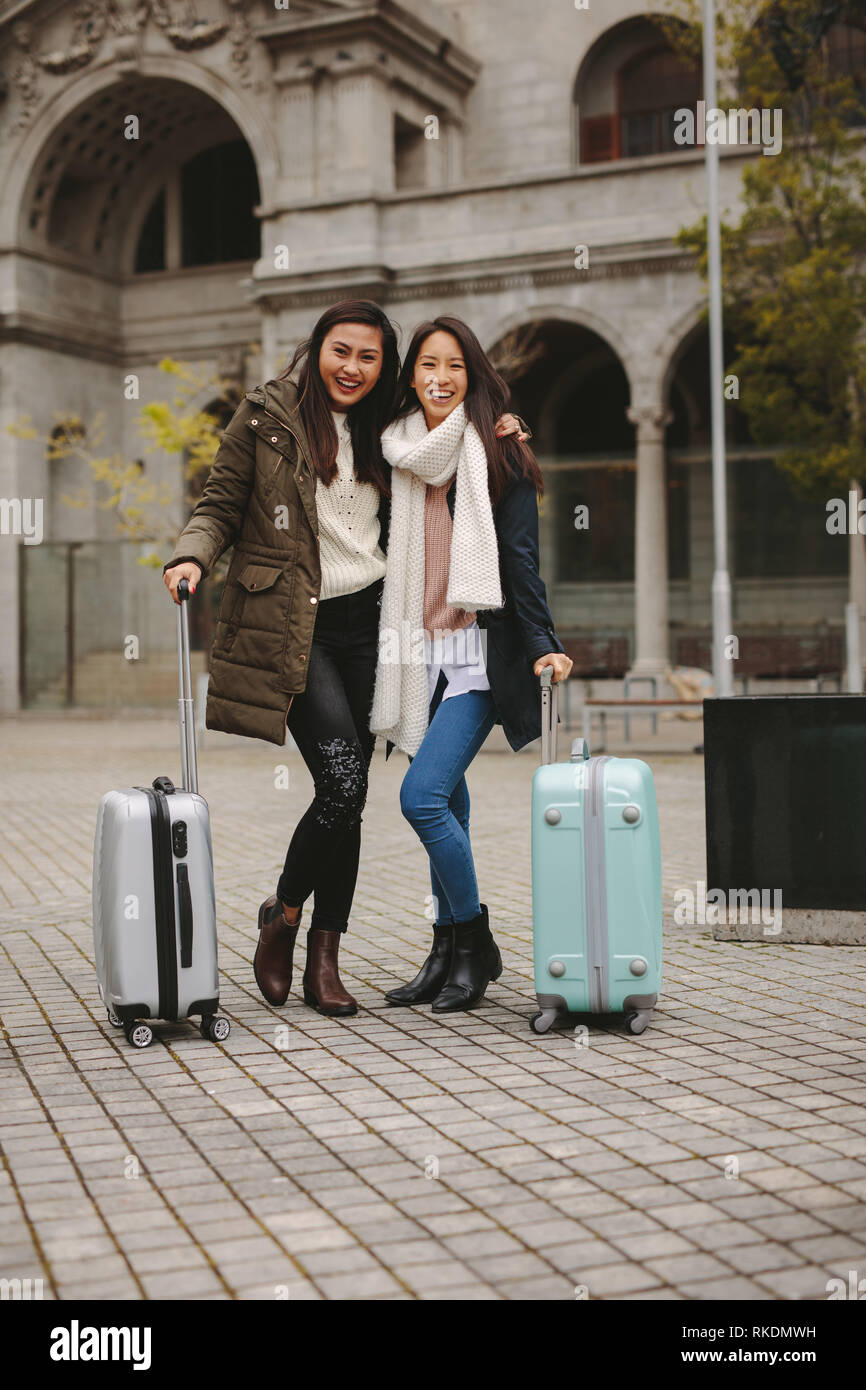 Two asian tourists standing in street holding their luggage bags. Cheerful female tourists in winter clothes going around the city. Stock Photo