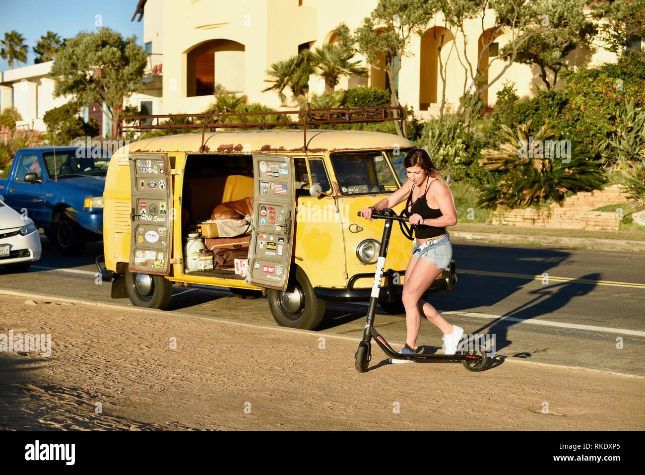 Attractive young woman hopping on Bird electric scooter next to classic Volkswagon VW minibus camper van parked at Sunset Cliffs, San Diego, USA Stock Photo