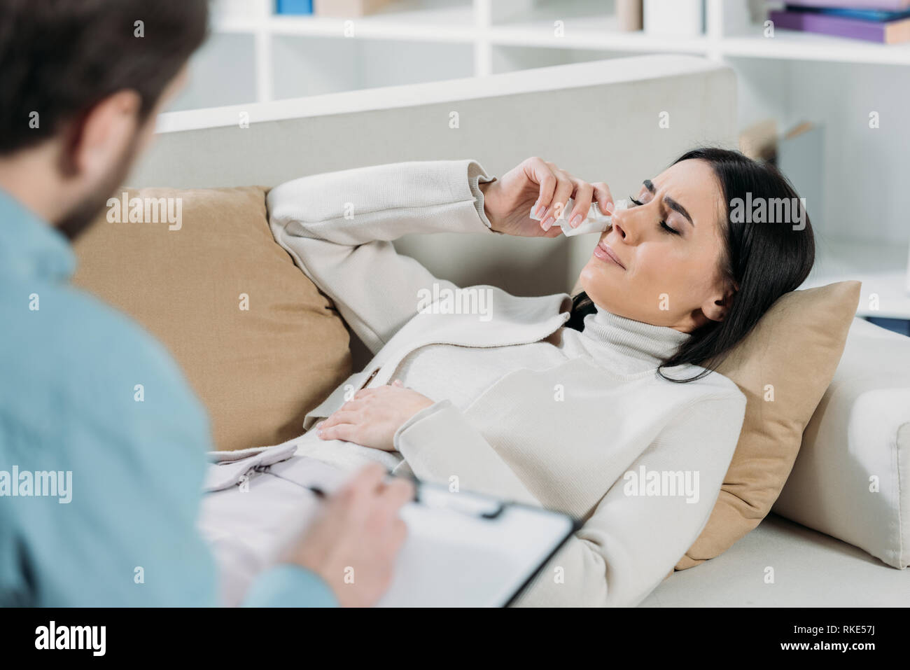 selective focus of psychotherapist writing on clipboard and young patient crying on couch Stock Photo