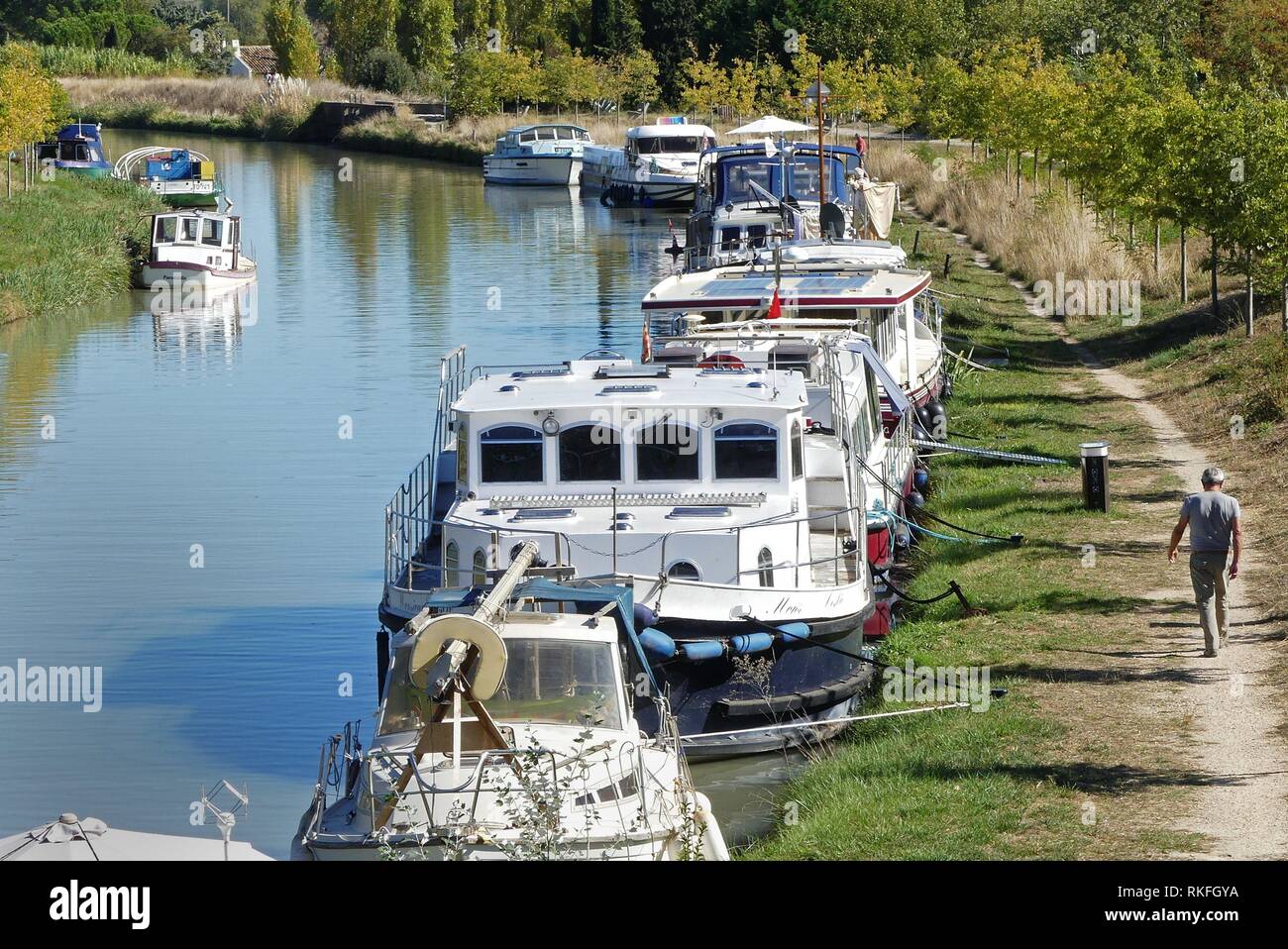 A man walks passed boats moored along the canal du Midi at Capestang, France Stock Photo