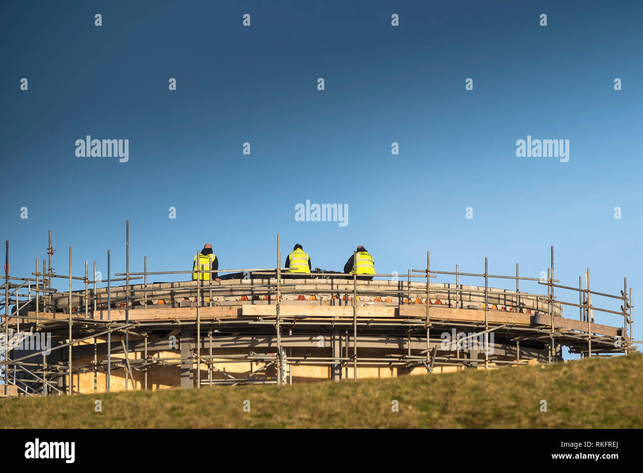Three construction workers wearing hi-viz jackets sitting on the wall of a structure surrounded by scaffolding. Stock Photo