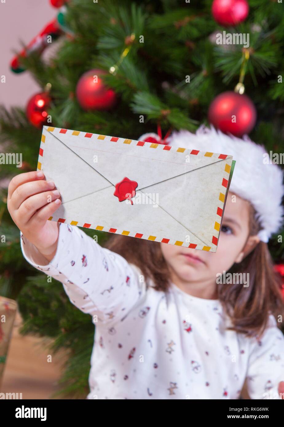 Little girl holds The Santa Letter Envelope. She is sitting beside Xmas Tree at home. Stock Photo
