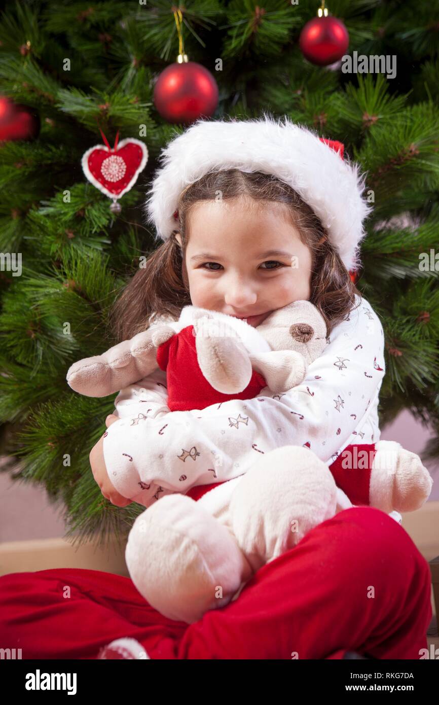Little child girl hugging her fluffy reindeer toy close to Christmas tree. She is very, very happy. Stock Photo