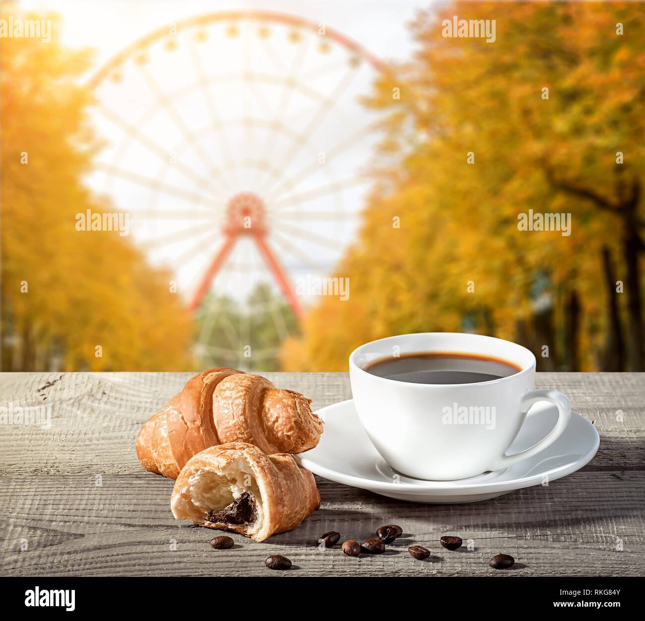 A cup of coffee with croissants on a wooden table. Grains of coffee on the table. Blurred background of a ferris wheel in autumn in the park. Stock Photo