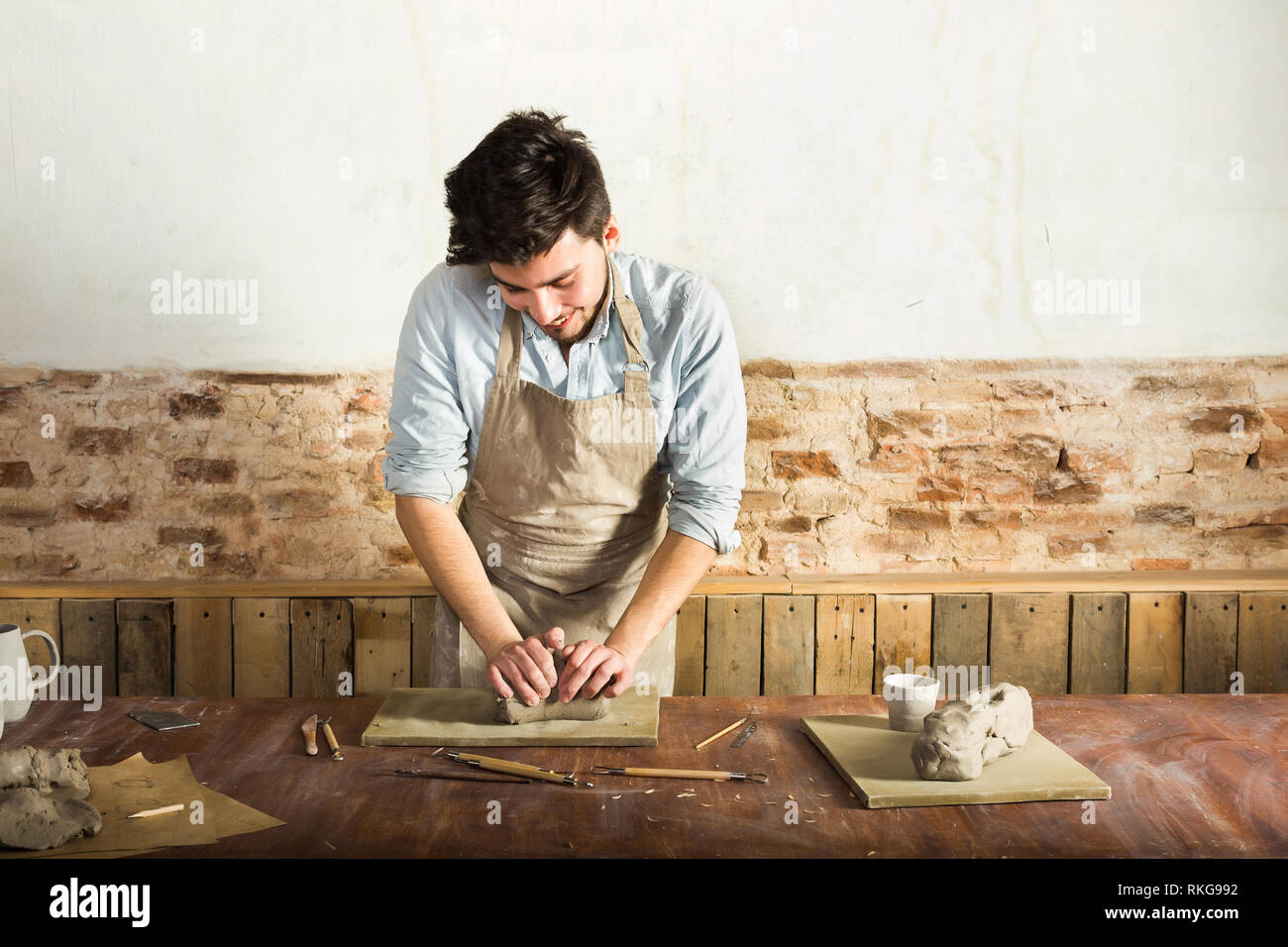 a young potter wearing apron and shirt kneading the clay on the desktop. Front view. Art and hobby and freelance working concept. Stock Photo