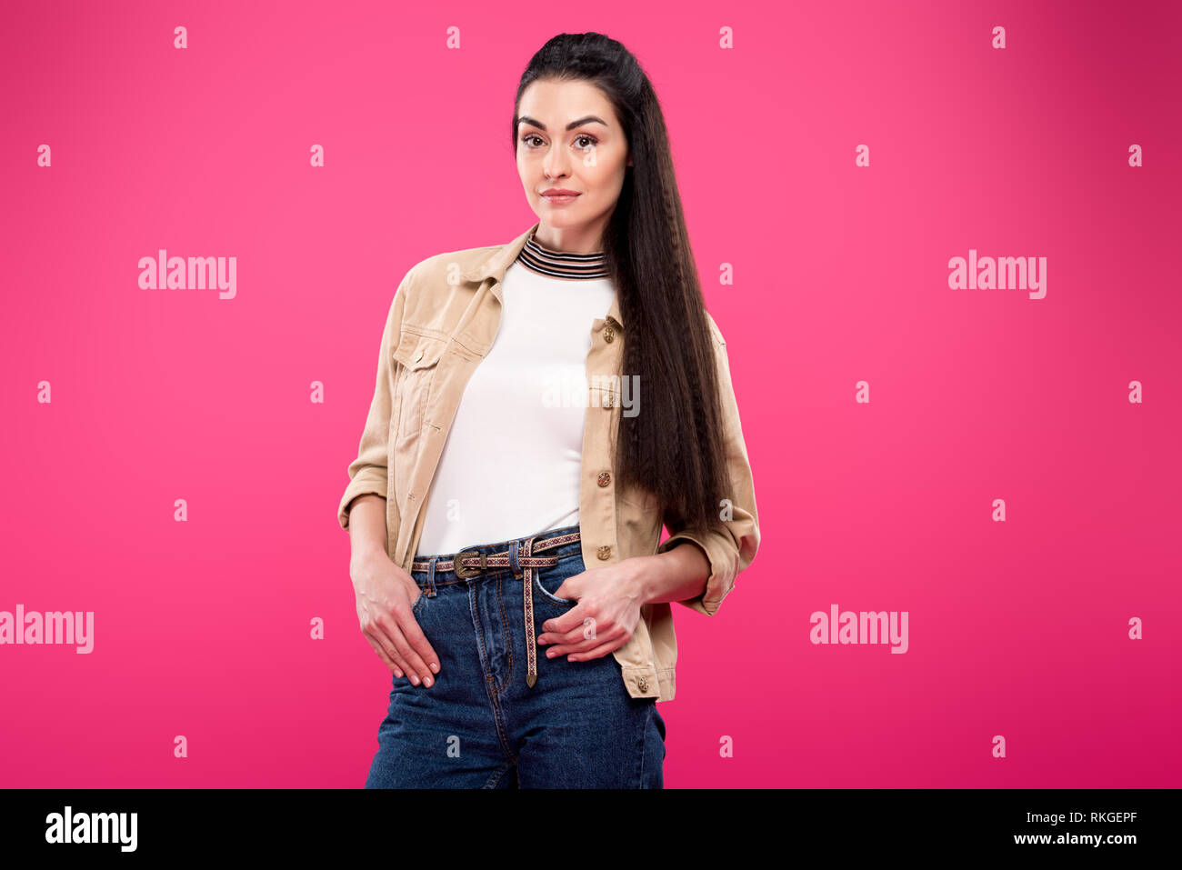 beautiful young woman standing with hands in pockets and looking at camera isolated on pink Stock Photo