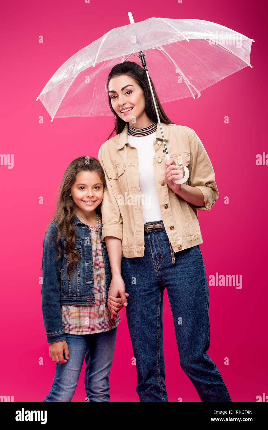 mother with umbrella and adorable happy daughter holding hands and smiling at camera isolated on pink Stock Photo