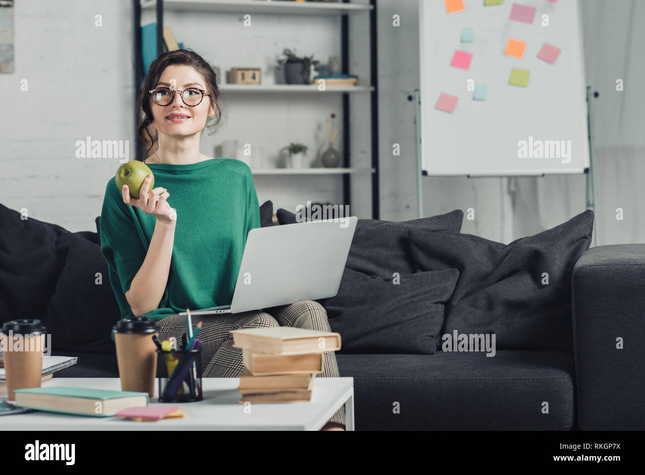 attractive woman holding apple and sitting on sofa with laptop Stock Photo