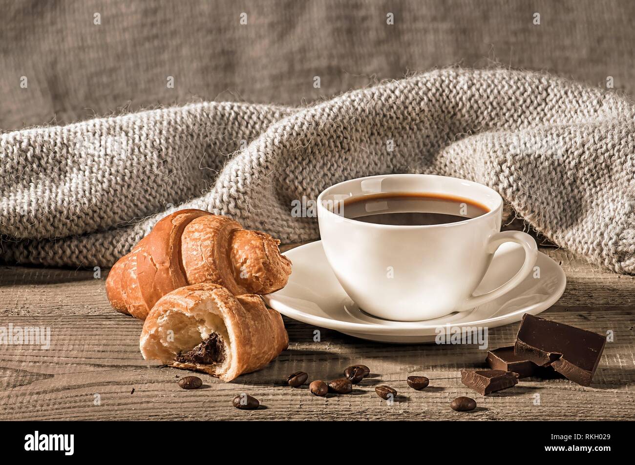 Coffee with croissants on the background of a woolen scarf. Grains of coffee next to the cup. Several pieces of dark chocolate next to each other. Stock Photo
