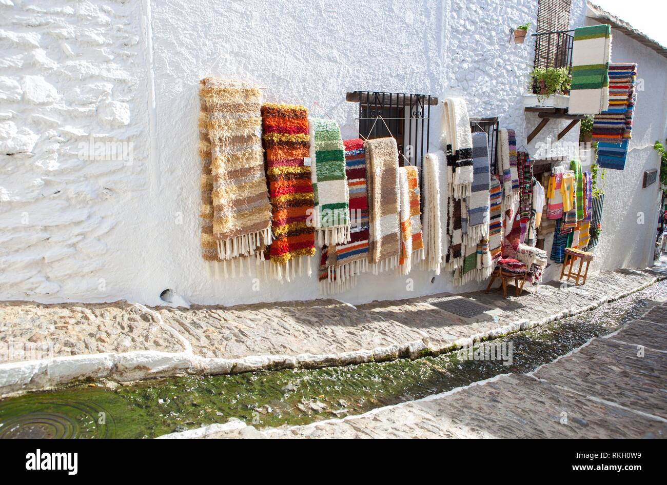 Jarapas for sell displayed over walls at quiet pretty street with water channel in the middle, Alpujarras, Granada, Spain. Stock Photo